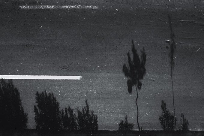 A grainy black-and-white nighttime photograph shows silhouettes of trees and foliage against a dark sky with two prominent horizontal light streaks, one near the top and another across the middle of the frame.