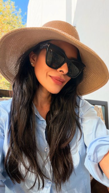 A woman takes a sunlit selfie wearing a wide-brimmed tan sun hat, dark sunglasses, and a light blue button-up shirt, her soft smile and relaxed pose capturing the cheerful, optimistic spirit of a happy Friday morning greeting.