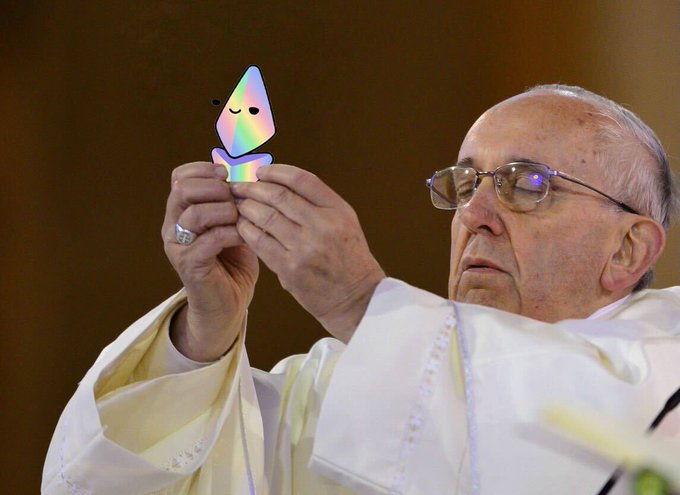 Pope Francis holds up a small, iridescent, hooded figure with a smiling face, examining it closely while wearing his traditional white vestments.