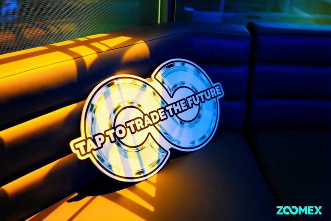 A stylized animated graphic shows two glowing, coin-like rings—an orange-gold ring overlapping a blue ring—on a dark blue couch or padded seat, with bold text across them reading “TAP TO TRADE THE FUTURE.” A small “ZOOMEX” watermark appears in the bottom-right corner.