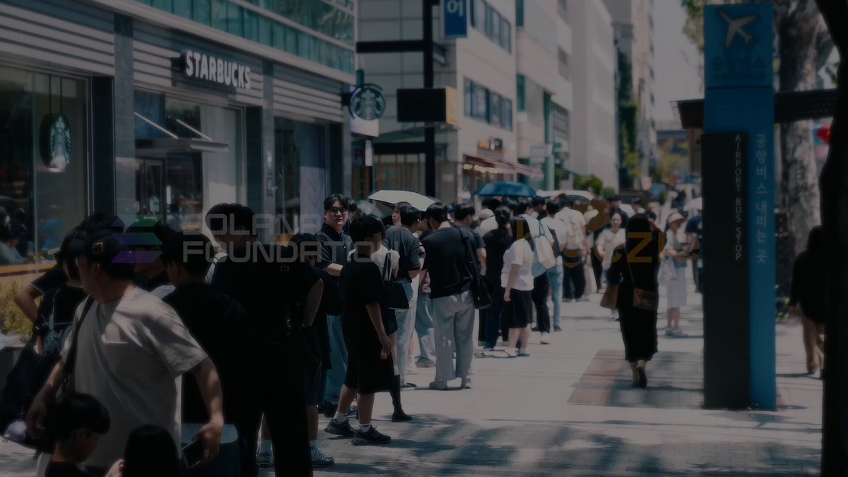 The image shows a large group of people standing outside Starbucks, waiting in line. The crowd includes individuals with backpacks and handbags, indicating that they might be tourists or travelers. There are also some umbrellas present among the crowd, suggesting that it could be a rainy day. The photo captures the atmosphere of a busy city street where people gather for various reasons such as shopping, dining out, or simply enjoying their time together.