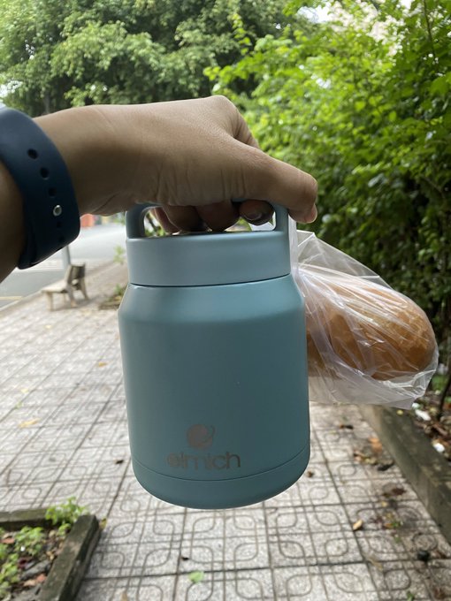 A hand holds a teal 'elmich' insulated food container with a clear plastic bag of bread buns beside it, against a backdrop of a sidewalk and green trees.