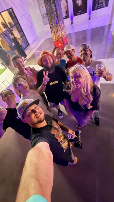A group selfie taken at what appears to be a party or gallery event, with several people posing together including a man in a straw hat making a peace sign, and colorful artwork visible in the background.