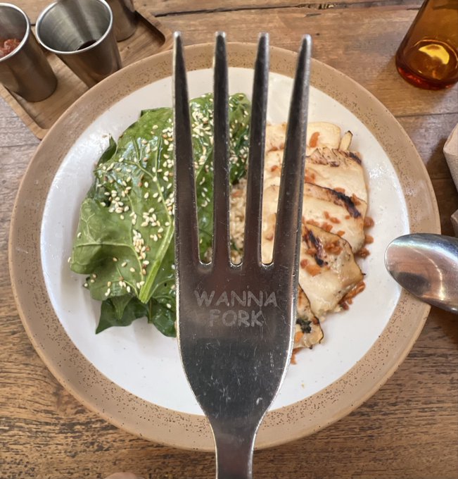 A close-up photo of a metal fork engraved with 'WANNA FORK' placed over a plate containing a spinach salad with sesame seeds and grilled chicken.