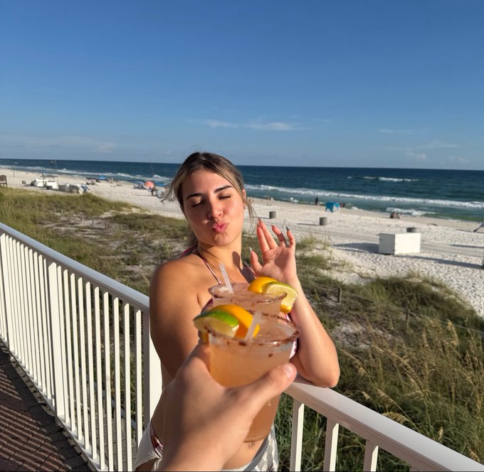 A young woman in a white bikini playfully puckers her lips on a balcony overlooking a sandy beach and ocean, while a hand in the foreground holds up a margarita glass garnished with lemon and lime.