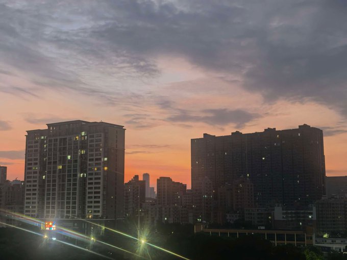 A dramatic sunset paints orange and purple hues across an urban skyline of high-rise residential buildings, with illuminated windows and light streaks from traffic below. The image evokes the contemplative, risk-focused tone of the post, visually representing a moment of pause to reassess crypto holdings (3 BNB at $710 avg and 3 OKB at $112 avg) against a backdrop that symbolizes the transition from one market phase to another.