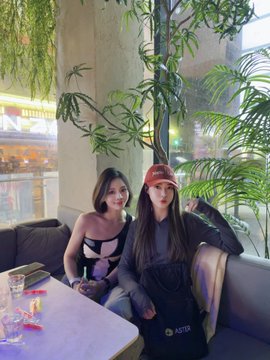 Two young women pose for a photo while sitting on a couch in a cafe or lounge area with plants in the background.