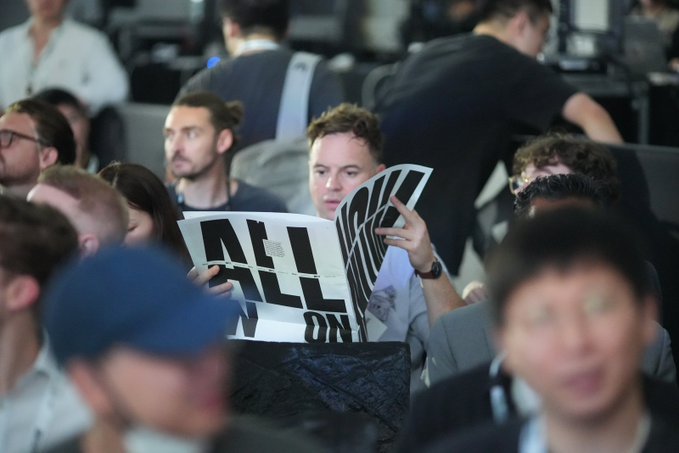 A man in a crowd holds up a black and white sign reading "ALL EYES ON..." at what appears to be a tech event or conference.