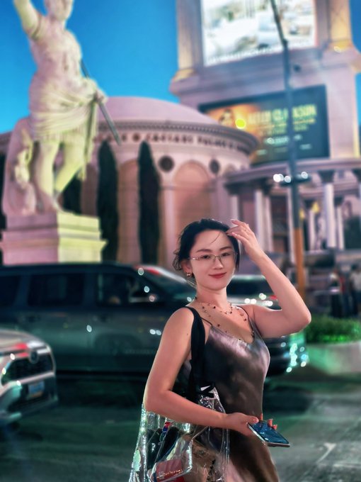 A young woman poses for a photo at dusk in front of a replica of Caesar's Palace and its statue in Las Vegas, holding shopping bags and a phone, with partial text "Caesar Club" visible on a sign behind her.