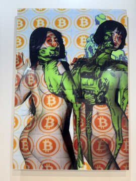 The image features two naked women with green faces, one wearing a mask and the other not. They are standing next to each other against an orange background adorned with circles of varying sizes. The caption below the image reads "Bitcoin is art - at @BTCPrague". This suggests that the artwork may be related to Bitcoin or cryptocurrency in some way, possibly as a commentary on its perceived artistic value or as a representation of its digital currency nature.