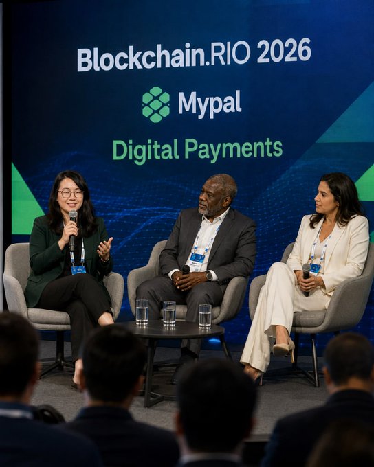Three panelists sit on stage at a conference with backdrop text 'Blockchain.RIO 2026', 'Mypal', and 'Digital Payments'. A woman on the left speaks into a microphone while a man and woman listen.