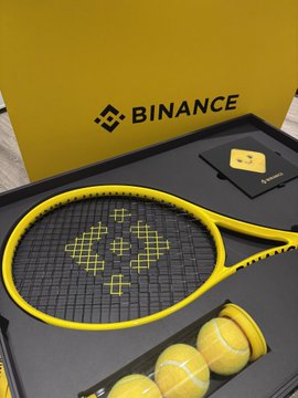 A yellow Binance-branded tennis racket and tennis balls are displayed in a black case with the Binance logo in the background.