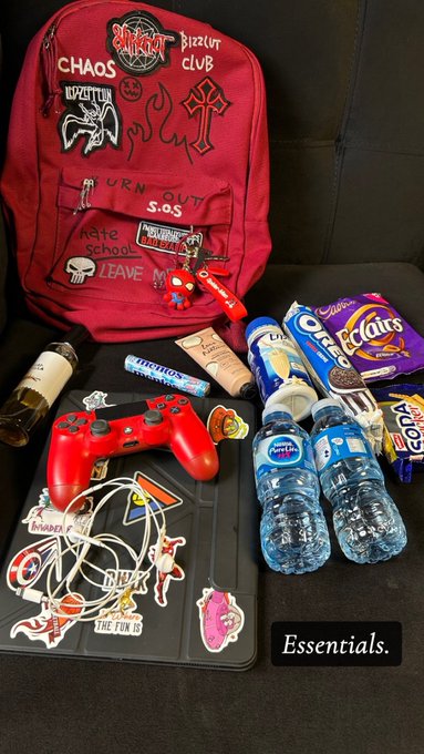 A red backpack adorned with patches reading "CHAOS," "Pizz Hut Club," and "Slipknot" sits alongside essentials like water bottles, Oreos, Eclipse gum, Smartfood popcorn, Airheads, and a Nintendo Switch controller, with the caption "Essentials." tying the scene to a "GM CT HAVE A BLESSED TUESDAY" greeting by showcasing items ready for a day out.