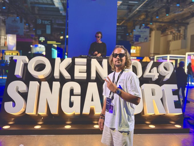 A man in sunglasses and a white t-shirt poses with a peace sign in front of a large illuminated "TOKEN 2049 SINGAPORE" sign at the crypto conference, while a DJ performs on a stage behind him, tying into the post's excitement about the upcoming "token Singapore" event.