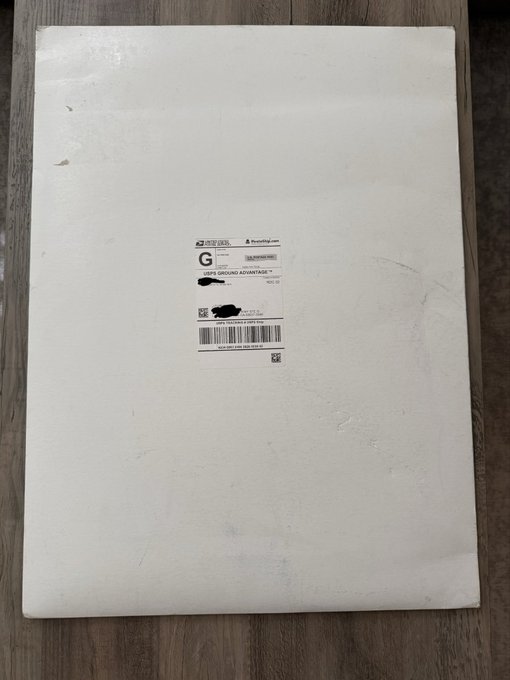 A white padded shipping envelope with a visible USPS shipping label, partially blacked out, lies on a wooden floor.
