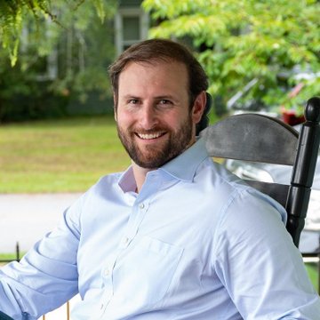 A smiling bearded man in a light blue button-down shirt sits outdoors, appearing relaxed and confident. His warm, approachable demeanor aligns with the caption quoting Patrick Witt expressing optimism about progress on the crypto CLARITY Act.