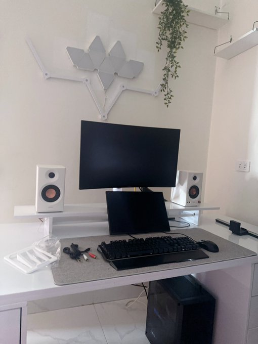 A minimalist white computer desk setup featuring a monitor, laptop, two white speakers with red cones, a gray desk mat, and geometric light panels on the wall above.