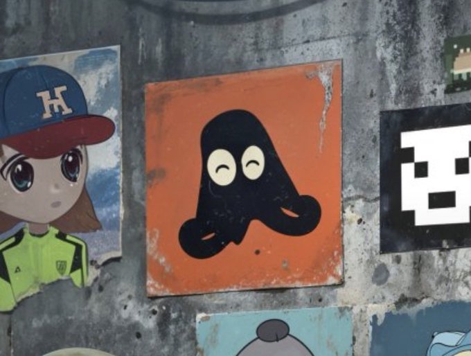 A collection of graffiti-style street art and posters on a wall, including a cartoon girl in a red and blue baseball cap and a black character with eyes on an orange background.
