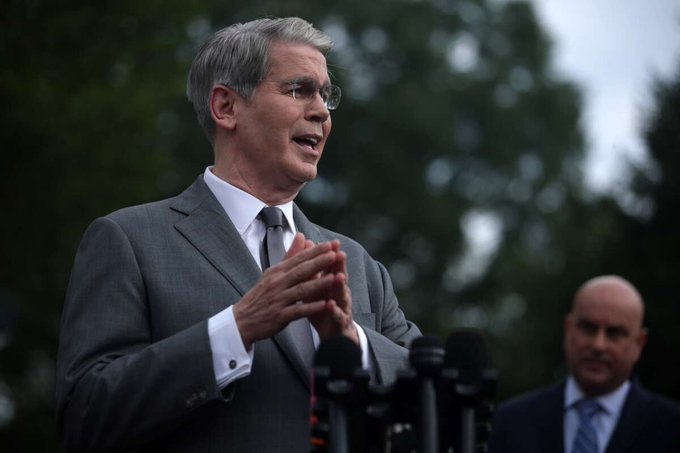 A man with gray hair and glasses wearing a gray suit speaks at an outdoor press conference with multiple microphones, with another man in a suit visible in the background.