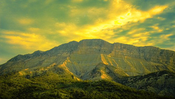 A dramatic mountain landscape under a vibrant yellow and green sky at sunset or sunrise, with rugged rocky peaks rising above a forested foreground.