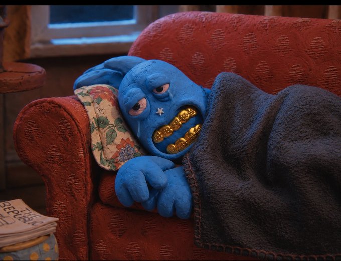 A sad blue claymation character with gold teeth lies on a red couch under a blanket, looking exhausted next to a newspaper.