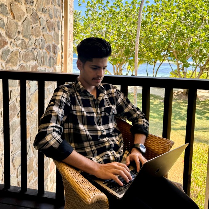 A young man wearing a checkered shirt works on a laptop while seated in a wicker chair on a balcony overlooking a tropical beach, illustrating the post's theme of embracing unconventional work hours to achieve trading freedom and flexibility in crypto.