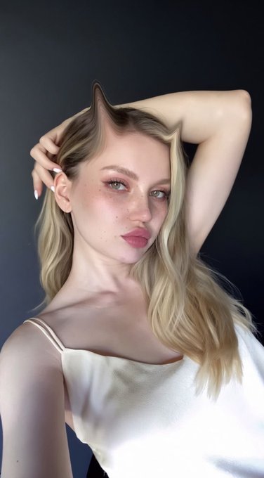 A young blonde woman with wavy hair poses for a portrait against a dark background, wearing a white satin top with her arm raised behind her head.