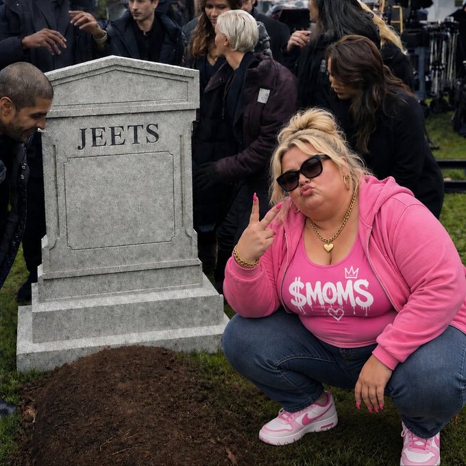 A woman wearing a pink hoodie, sunglasses, and a '$MOMS' t-shirt crouches and poses with her middle finger raised next to a tombstone labeled 'JEETS' at what appears to be a filming set.