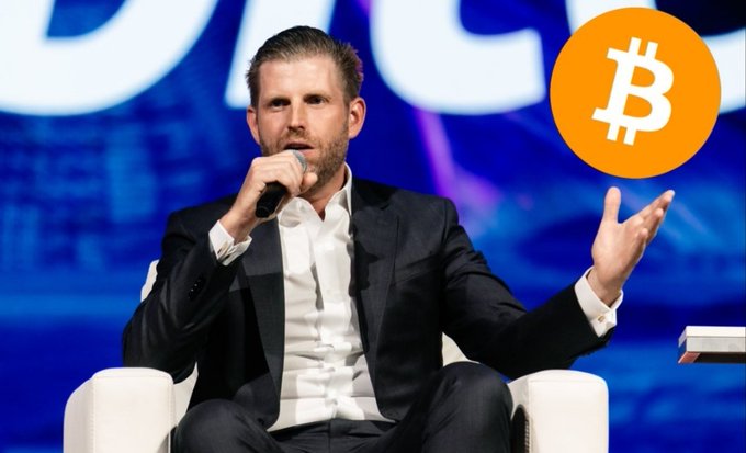 Eric Trump speaks into a microphone at what appears to be a conference, with a Bitcoin logo displayed prominently in the upper right corner. The image illustrates the accompanying post about American Bitcoin, an Eric Trump-backed company that recently expanded its treasury to 8,300 BTC through an additional 300 Bitcoin purchase.