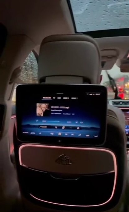 A rear-seat entertainment screen mounted on the back of a Maybach headrest displays a music player interface, with rain visible on the panoramic sunroof above and red taillights glowing through the rear windshield. The luxurious cabin setting reflects the post's theme of grinding after leaving a Web2 job, suggesting the speaker has leveled up to a lifestyle where they're chauffeured while monitoring crypto's chop around $65k and the AI rotation.