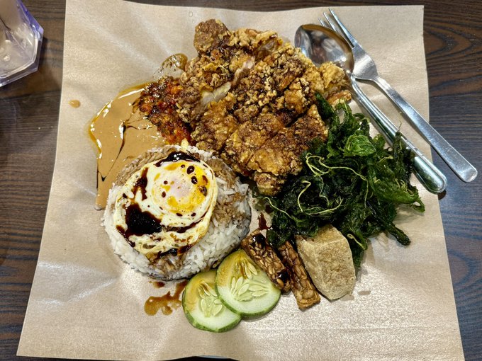 A plated meal served on parchment paper featuring rice topped with a fried egg, fried chicken, leafy greens, cucumber slices, and sauces, with a spoon and fork resting on the side.