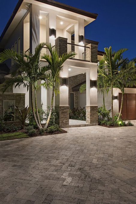 A nighttime photo of a modern, luxury two-story house with palm trees, stone accents, and outdoor lighting illuminating the entrance and driveway.