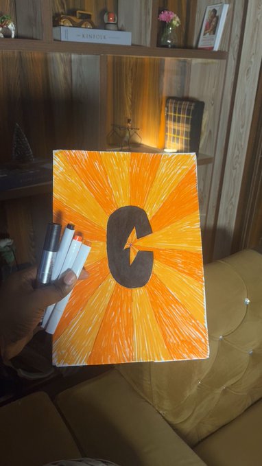 A hand holding marker pens next to a hand-drawn artwork featuring a dark letter 'C' with a lightning bolt cutout on a radiating orange and yellow sunburst background, displayed in a living room setting.