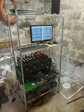 A makeshift cryptocurrency mining rig built on a wire shelving unit, featuring eight GPU cards stacked below a monitor displaying system metrics, with two cooling fans placed on the floor to manage heat. The setup illustrates the escalating hardware costs referenced in the post, highlighting the challenge of competing with industrial-scale mining operations.