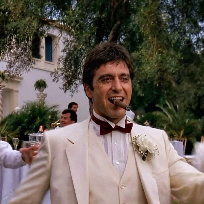 A man in a cream-colored tuxedo with a burgundy bow tie and a white boutonniere, smiling broadly with a cigar in his mouth at an outdoor celebration with greenery in the background.
