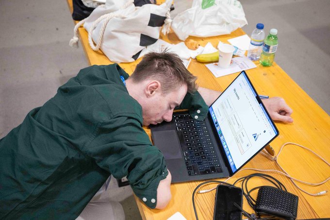 A young man in a green shirt has fallen asleep with his head resting on a MacBook laptop on a wooden table, surrounded by water bottles, a banana, snack wrappers, and tangled cables—a classic scene of exhaustion following a hackathon.