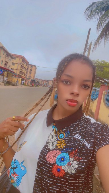 A woman with braided hair wearing a floral patterned shirt takes a selfie on a street lined with buildings and palm trees.