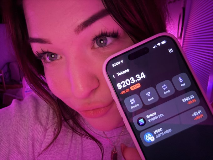 Close-up of a young woman with long lashes bathed in pink/purple light, holding up a smartphone showing a crypto wallet app with $203.34 balance, including Solana and USDC holdings.