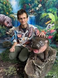 A man kneeling in a jungle setting holding a baby hippopotamus, with other hippos visible in the background.