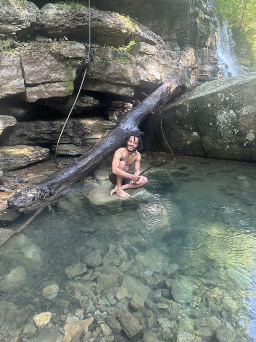 A young man crouches on a rock at the edge of a clear, rocky stream beside a fallen log and a small waterfall.