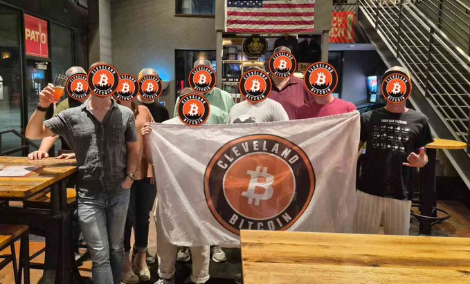 A group of people whose faces are obscured by 'Cleveland Bitcoin' logos holding a matching banner inside a bar with an American flag visible in the background.