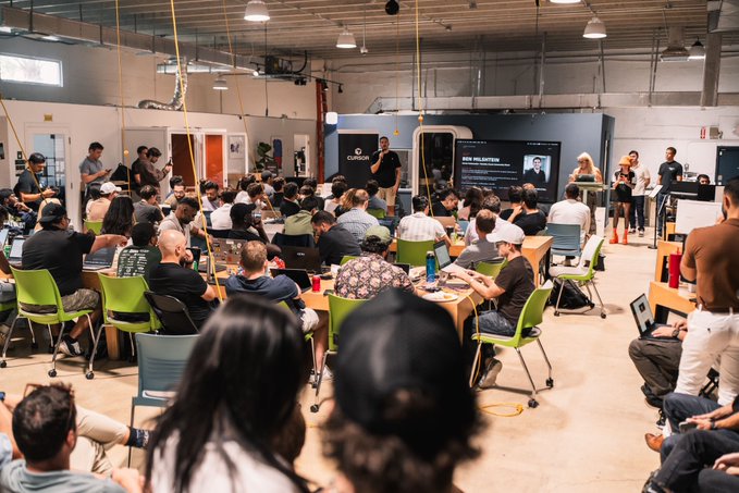A large group of people seated in a workshop or conference setting in an industrial-style venue, with a presentation happening at the front.