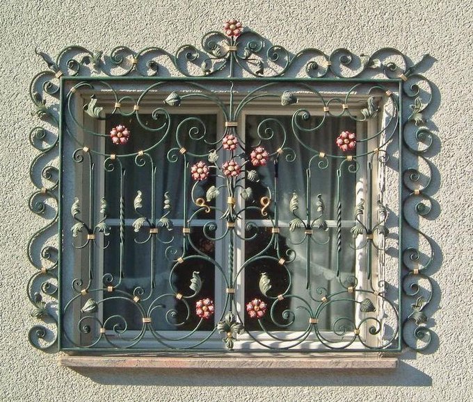 An ornate wrought iron window grate features intricate scrollwork and decorative floral motifs in red and gold, mounted on a textured cream-colored wall. Behind the decorative metalwork, a window with sheer curtains is visible, allowing soft natural light to filter through.