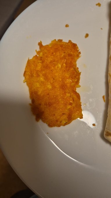 A close-up photo shows a golden-brown, crispy breaded cutlet or hash brown patty resting on a white plate, with crumbs scattered around it.