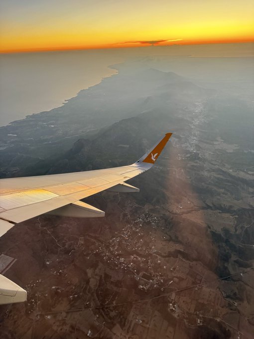 An aerial view from an airplane window showing the wing at sunset, with the orange winglet bearing a logo, overlooking mountains and coastline.