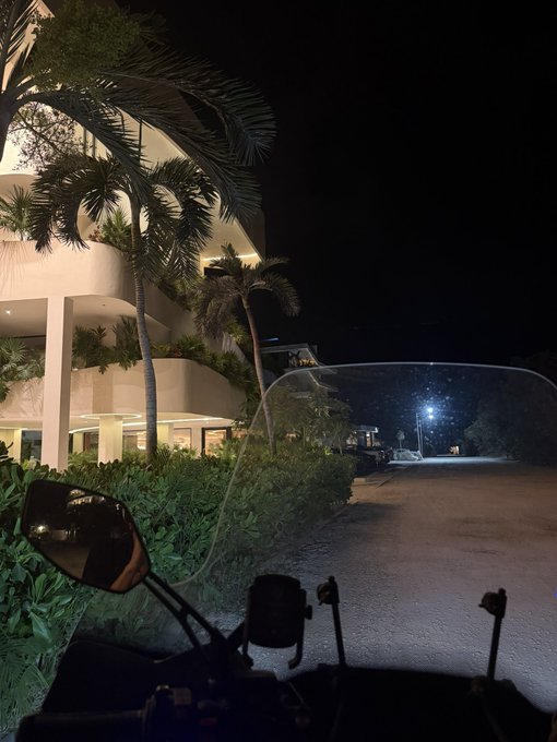 A nighttime view from a motorcycle, looking past the windshield and side mirror toward a lit building with palm trees and an empty road.