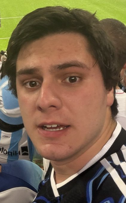 A close-up selfie of a young man with dark hair wearing a blue and black sports jersey, with another player visible in the background on a green field.