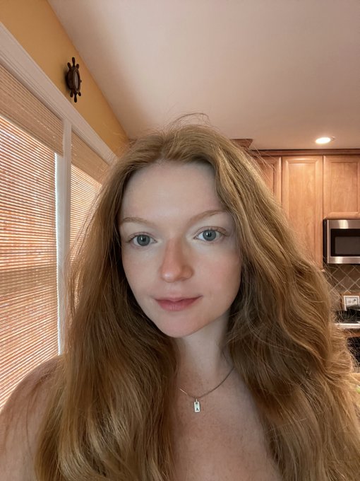 A selfie of a young woman with long red hair in a kitchen setting, wearing a small pendant necklace.