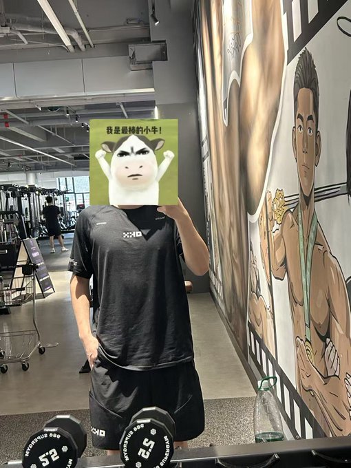 A person in workout attire holding a green card with a cat face and Chinese text '我是最棒的小牛!' in a gym setting.
