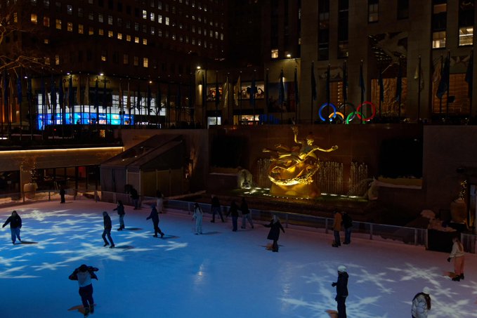 An outdoor ice skating rink at night illuminated by blue star projections, with a golden Prometheus statue and Olympic rings visible in the background at Rockefeller Center.